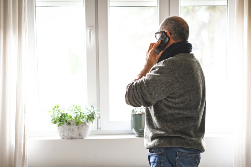 Why Won’t My Furnace Stop Running? Photo of an elderly man talking on the phone while looking out of a window, seemingly upset.